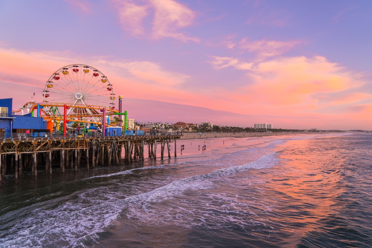 Santa Monica Pier featuring Pacific Park's Ferris wheel and roller coaster over the ocean during a pink and orange sunset.